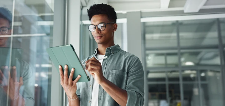 Ricoh Process Automation - A person holding a green tablet and looking down at it. They are wearing glasses and a light-colored shirt. The background is a glass window and a blurred office space.